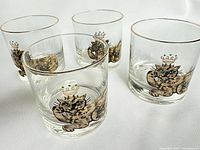Four clear lowball glasses with a gold rim, each featuring a gold cat with green eyes and a crown design by Couroc.