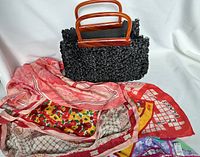 Black woven raffia handbag with tortoise-shell style handles displayed with four folded silk scarves in various colors and patterns including floral and geometric designs.