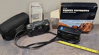 Two vintage 35mm film cameras - The Nature Company Panorama Camera and Ricoh Zoom S - with their boxes and a black soft camera bag placed on a surface, measuring tape showing size.