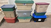 9 Rubbermaid storage totes stacked on driveway in various colors and sizes with lids, some with wheels.