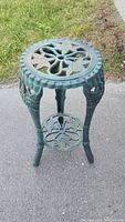 Full view of green cast iron plant stand on pavement with visible intricate floral design on top tier and legs.