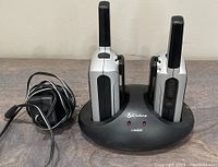 Two Cobra MicroTalk radios placed in the black dual charging stand with power adapter cable coiled beside it.
