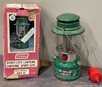 Overall view of the Coleman Sport-Lite Lantern Model 321 C with the original box. Shows the lantern standing upright with a red plastic cap accessory beside it and the colorful, worn box in the background.