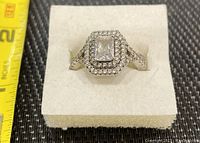Front view of the sterling silver ring with central rectangular clear stone, double halo of small stones, and textured band with small stones.