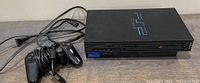 Sony PlayStation 2 slim console with one black wired controller lying on top, controller cable coiled.