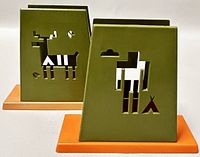 Two vintage St. Labre Indian School plastic napkin holders with green trapezoidal bodies and cutout horse, cloud and teepee designs mounted on orange and mustard yellow bases.
