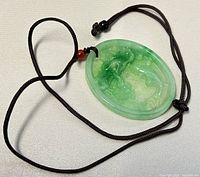 Close up of the oval green jade pendant with visible detailed carvings and attached adjustable black cord with a red bead accent.