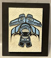Full front view of the embossed plaque showing the indigenous style artwork mounted on textured cream background panel framed by dark brown wood.