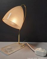 Mid-century modern style Lucite and brass table lamp with conical shade lit up, viewed from the side against a black background.