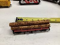 Model train flatcar loaded with two logs, shown with measuring tape indicating size around 7 inches long.