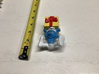 Smurf figurine lying on back holding a present, with measuring tape for scale