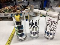 Three clear glass tumblers featuring different Beatles designs placed side by side on a table surface with a measuring tape nearby for scale.