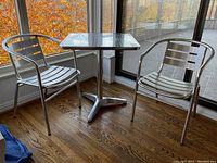 Patio set including square aluminum table and two aluminum chairs with slatted design, shown in indoor setting near window with fall foliage outside.