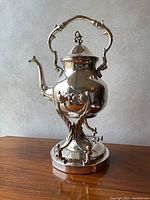 Front side view of the antique silver plated tilting teapot on its stand, showing handle, spout, and base.