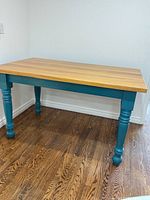 Three-quarter front view showing natural pine top and teal painted turned legs.