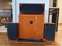 All four speakers arranged on floor, one center subwoofer front and center, two smaller speakers on sides, one center speaker on top of subwoofer showing wooden enclosures with visible wear and black fabric grills.
