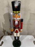 Smaller wooden nutcracker standing on flat surface with black hat, white feather, red gem, red and white uniform, black boots, and sword.