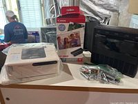 Canon Selphy printer on left, original box with cartridges in center, Panasonic charger on right with bag of batteries in front