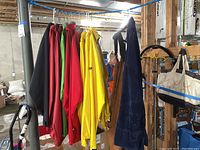Full rack of raincoats and foul weather gear hung on a clothes rack in a basement, showing multiple jackets mostly in black, red, green, yellow, plus a dark blue work overall.