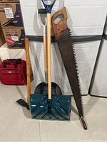 Photo showing full view of shovels, pickaxe, and large hand saw against white storage background