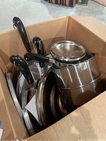 Box containing stainless steel pots with black handles, stacked and nested inside each other.