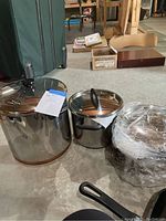 Three stainless steel sauce pots with lids shown on concrete floor. Pots have reflective surfaces and black lid knobs. IKEA griddle pan partially visible to the right.