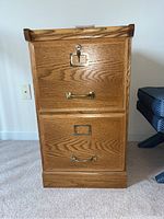Front view of oak two-drawer file cabinet showing grain pattern, brass hardware, label holder, and lock on top drawer with key inserted.
