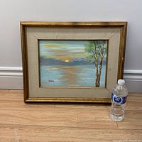 Frontal view of the signed oil on board landscape painting with gold frame and beige matting, showing the two trees to the right, lake or water body, mountains, and sunset/sunrise sky.