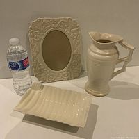 Photo of three cream-colored decorative pieces including a mouth blown glass frame with intricate pattern, a tall pitcher, and a wavy edged rectangular dish. Water bottle included for scale.