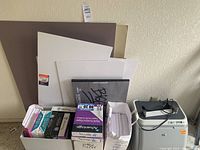 Photo showing multiple unopened and open boxes of copy paper, foam board, newsprint pad, and large poster boards stacked against the wall with HP laser printer cord nearby.