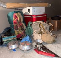 Photo showing sleeping bag, vintage Coleman ice chest, rolled wicker beach mats, outdoor books, canteen with cover, and cooking utensils