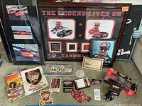 Wide view of various framed Dale Earnhardt memorabilia, collectible race car models, race programs, and assorted racing ephemera arranged on a table and visible in the background.