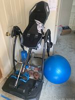 Full view of Teeter inversion table with attached footrests, blue foam roller next to it, blue exercise ball, metal dumbbell with weight plates resting near base.