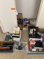 Overview photo showing several boxes filled with assorted office supplies including staplers, tape dispensers, clips, Post-it notes, and calculators.