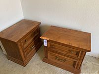 Pair of medium-toned oak wood nightstands with two drawers each. Brass handles and knobs. Positioned on carpet near a white wall.