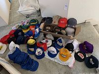 Photo of approximately 30 assorted snapback hats arranged on floor and in a box, showing wide variety of logos and colors including sports and company branding