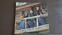 Front cover of America Hideaway vinyl album featuring three band members on cabin porch.