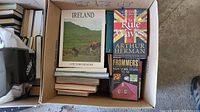 Top view of an open box containing multiple travel and sightseeing-related books including 'Ireland: A Picture Memory,' 'To Rule the Waves' by Arthur Herman, and 'Frommer's New York State 72-73' travel guide.
