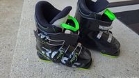 Pair of black Comp J3 ski boots with green details, showing left and right boots side by side on concrete surface.
