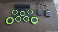 Photo showing electronic drum pad with seven green and black round playing surfaces and two wired foot pedals on a table.