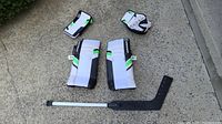 Full set of Warrior road hockey goalie equipment including leg pads, blocker, catcher glove, and goalie stick on concrete ground