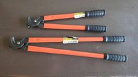 Photo showing two cable cutters with orange handles lying on a surface, showing the full length and cutting heads with labels visible.