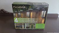 Box front showing brand Paradise and 4 solar LED accent lights mounted on fence posts.