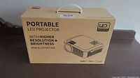 Front and side view of box showing an illustration of the portable LED projector and the text highlighting its features such as higher resolution and brightness, mini and ultrathin design.