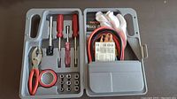 Top view of open gray plastic tool case showing hand tools, electrical tape, nuts, jumper cables, and gloves neatly stored inside.