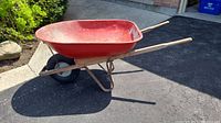 Side view of red metal wheelbarrow showing wooden handles and black tire with white rim.