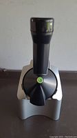 Front view of Yonanas frozen yogurt maker showing chute with black plunger and outlet feature with green swirl emblem.