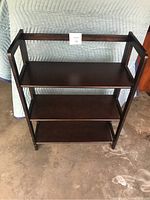 Front view of solid wood 3-tier folding bookshelf against padded backdrop on concrete floor.