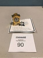 Miniature brass clock on a white surface with paper tags and listing number in image