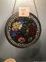 Round stained glass suncatcher hanging against a wall, showing the floral design with various colors including red, pink, blue, yellow, and green.